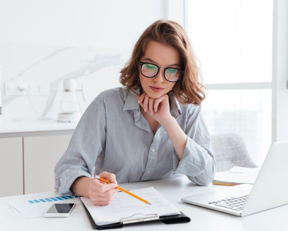 Young concentrated businesswoman in glasses and striped shirt working with papers at home